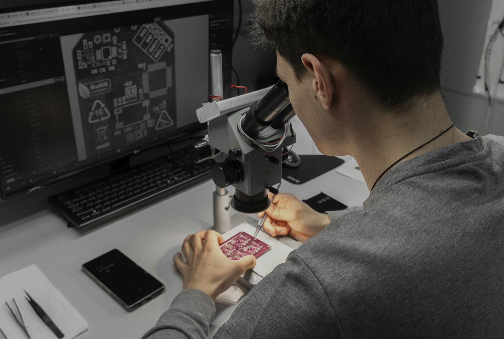 Person arbeitet am Mikroskop an einer Platine, Computerbildschirm im Hintergrund.